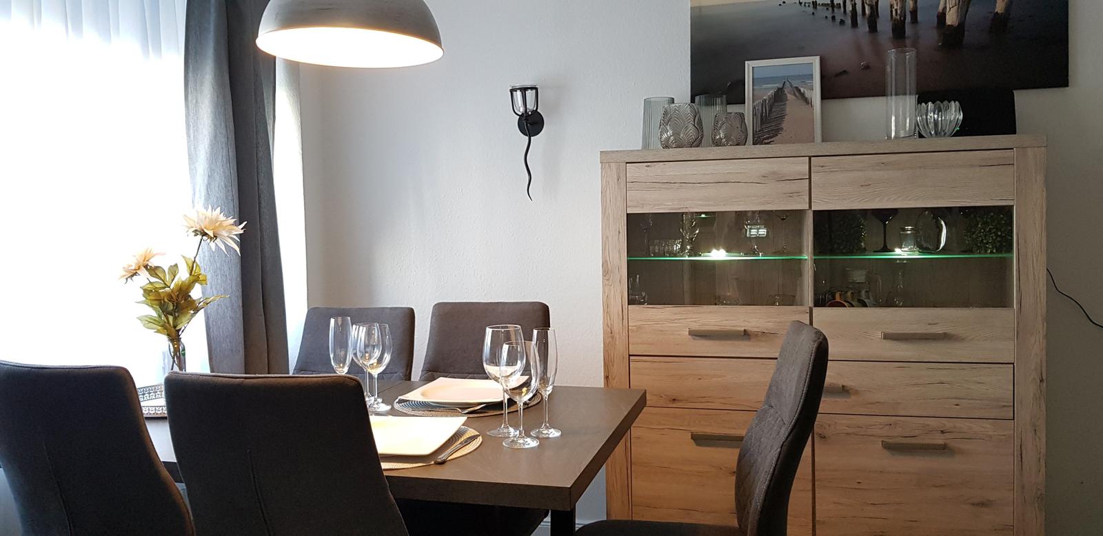 Esszimmer mit Tisch, Stühlen und Glasvitrine. Fenster mit Vorhängen und Wandbeleuchtung.