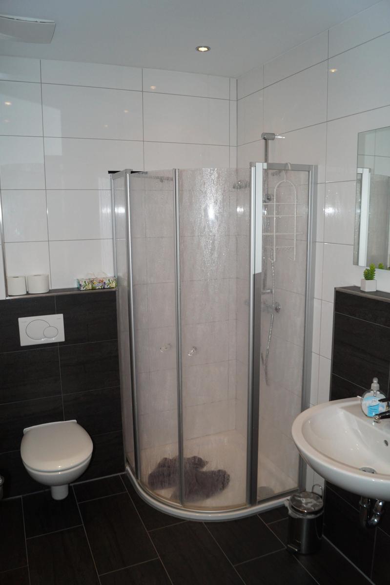 Badezimmer mit Dusche, Waschbecken und Toilette. Weiße Fliesen und dunkle Bodenfliesen.