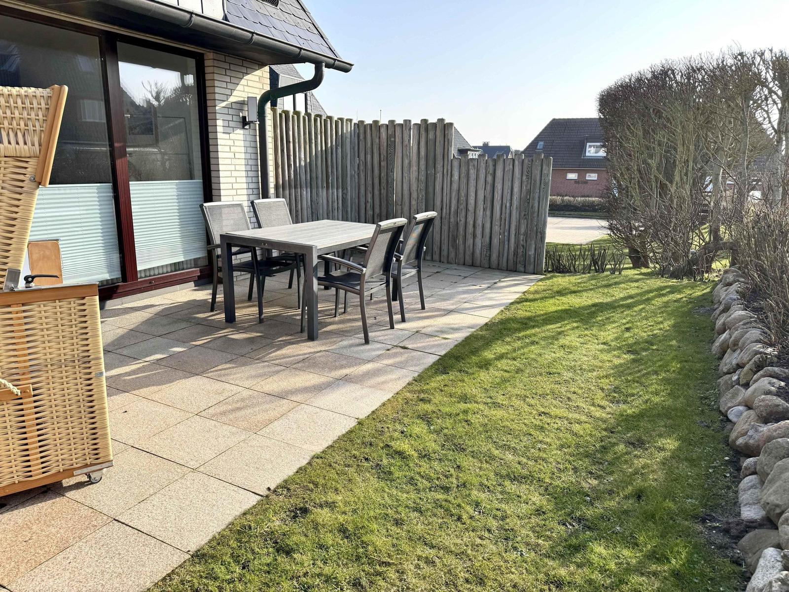 Terrasse mit Tisch und Stühlen, Grasfläche und Holzzaun. Wickermöbel links.