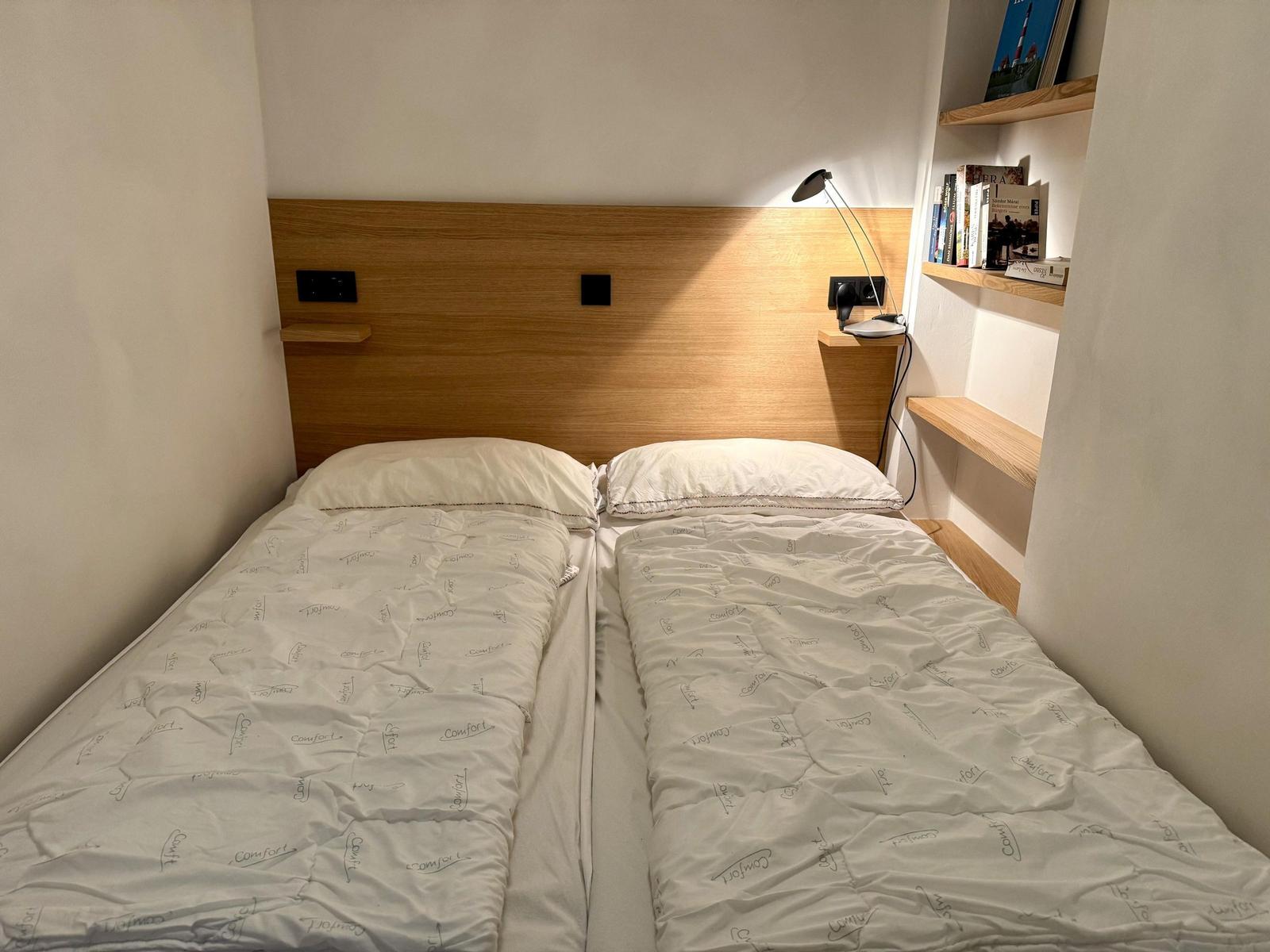Zwei Betten mit weißen Bettdecken und Holz-Kopfteil. An der Wand hängen Bücherregale.