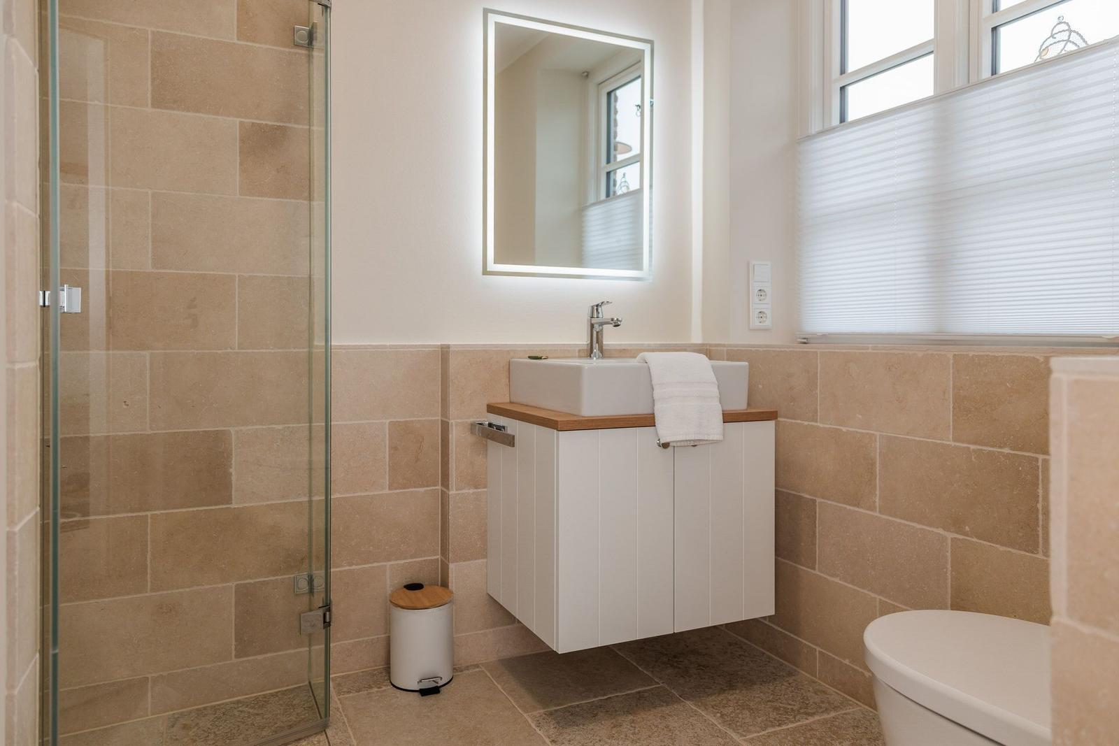 Badezimmer mit Dusche, Waschbecken und Toilette. Fliesenwand und Spiegel mit Beleuchtung.