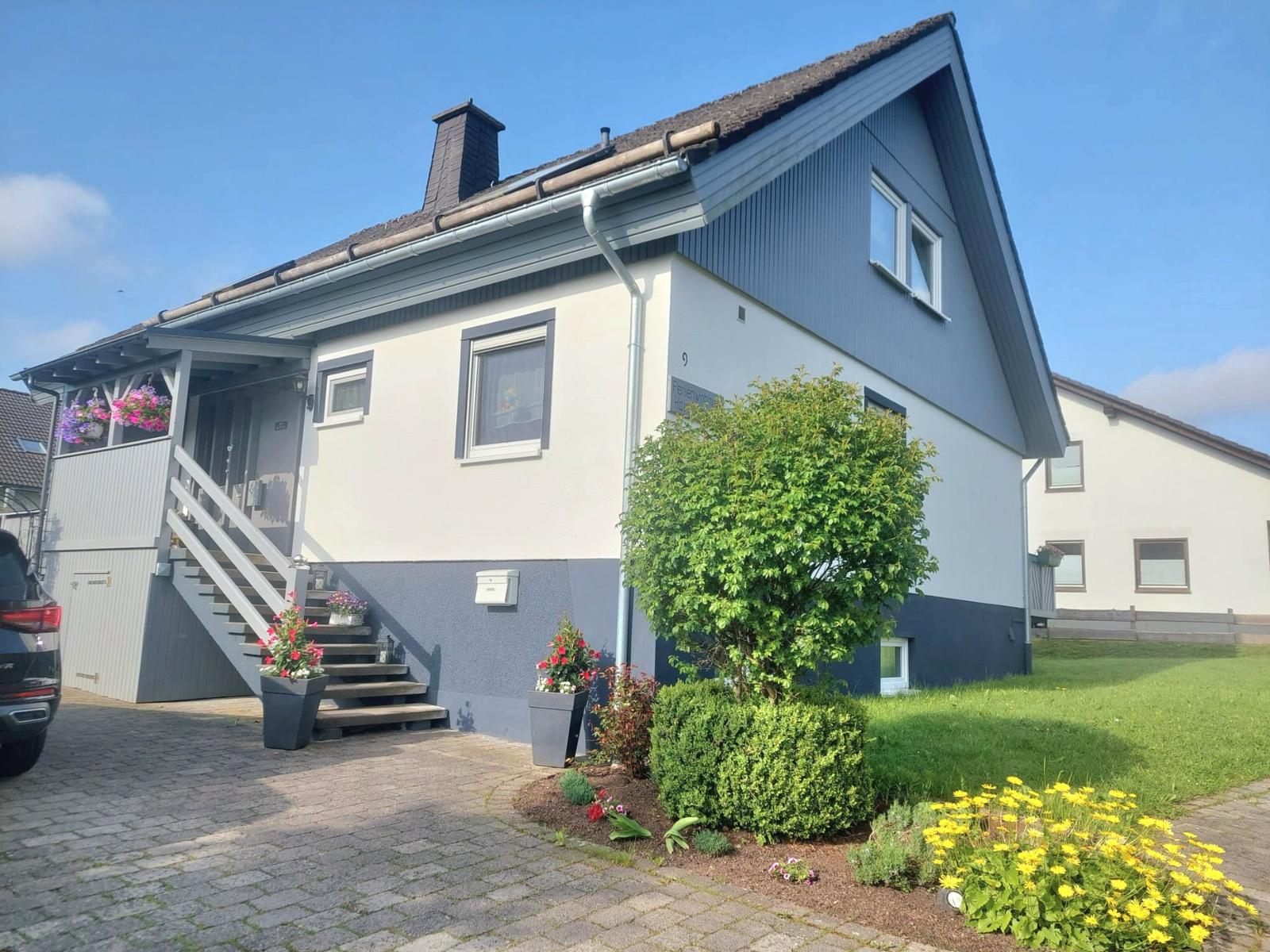Haus mit Eingangstreppe, Blumenbeete und gepflastertem Vorplatz unter blauem Himmel.