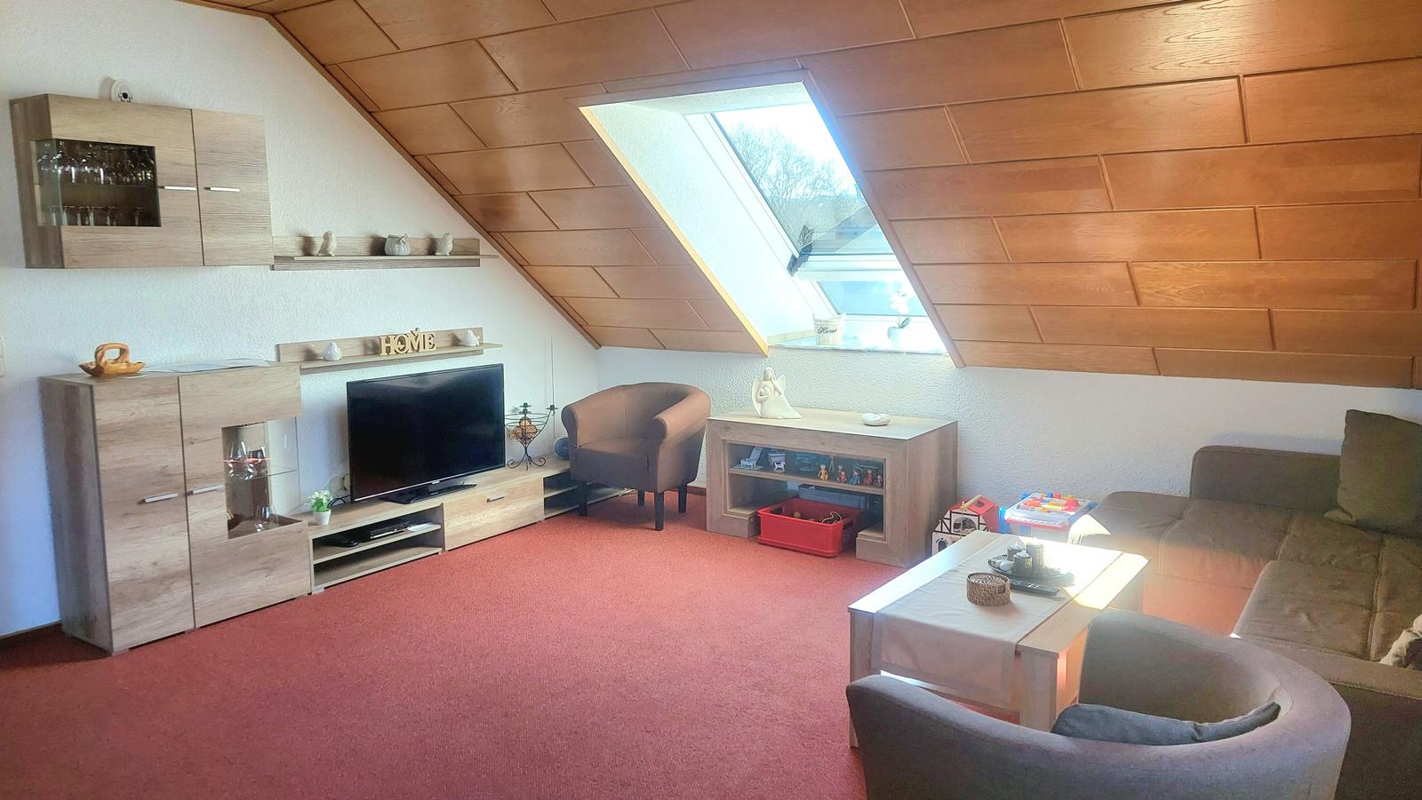 Wohnzimmer mit Holzdecke, Fernseher, Sofa und Sessel. Fenster mit Blick nach draußen.