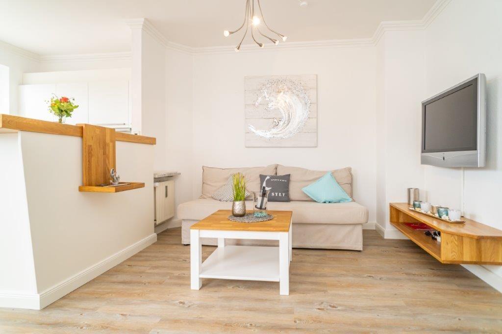 Wohnzimmer mit Sofa, Couchtisch und Wand-TV. Holzfußboden und Küchenbereich sichtbar.