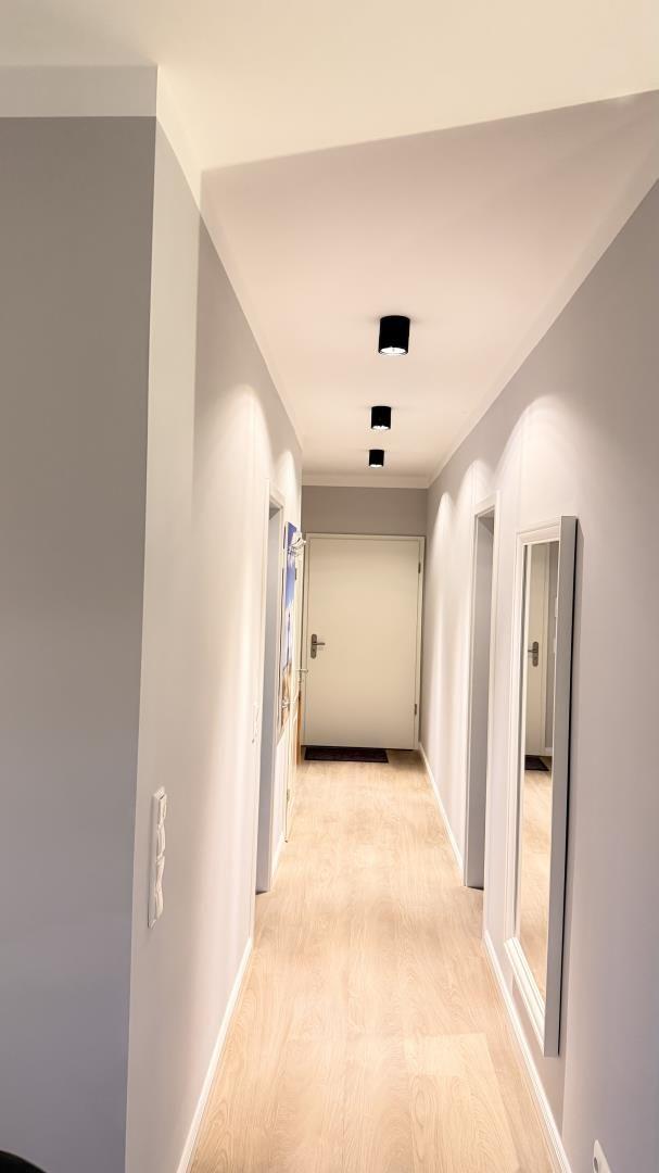 Flur mit hellen Wänden, Holzboden und Deckenleuchten. Türen zu verschiedenen Räumen sind sichtbar.