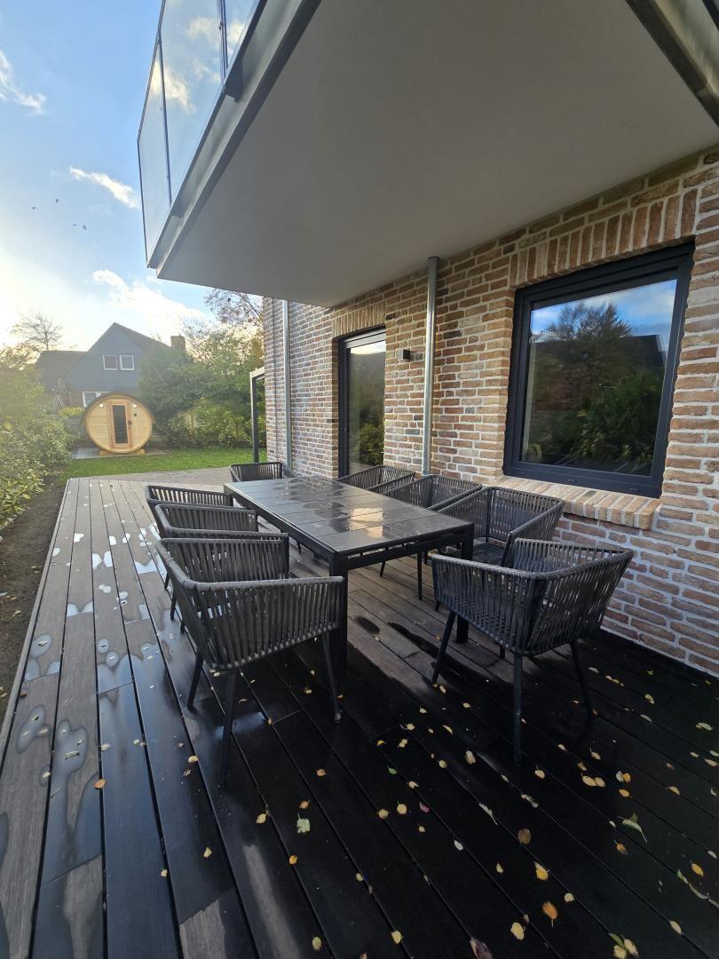 Terrasse mit Tisch und Stühlen, dunkler Holzboden, Backsteinwand und großes Fenster.