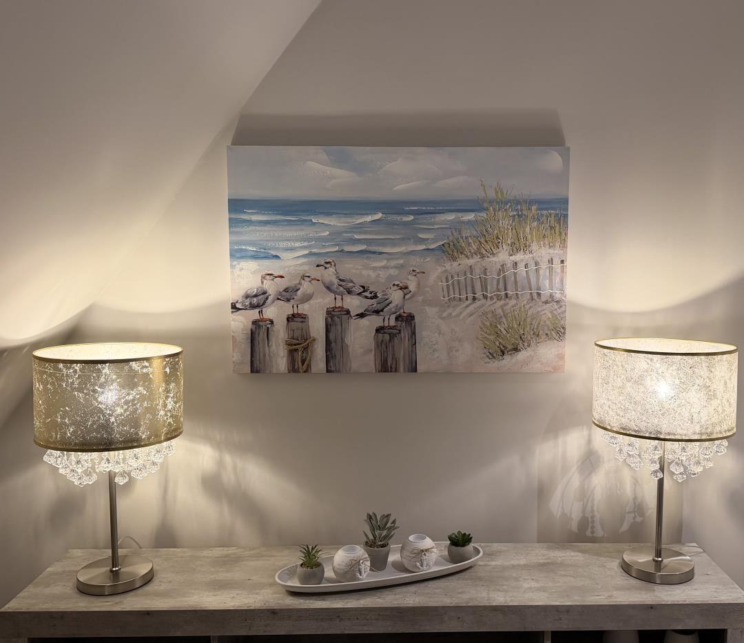 Wandbild mit Strandmotiv über einem Tisch mit zwei Lampen und Pflanzen.