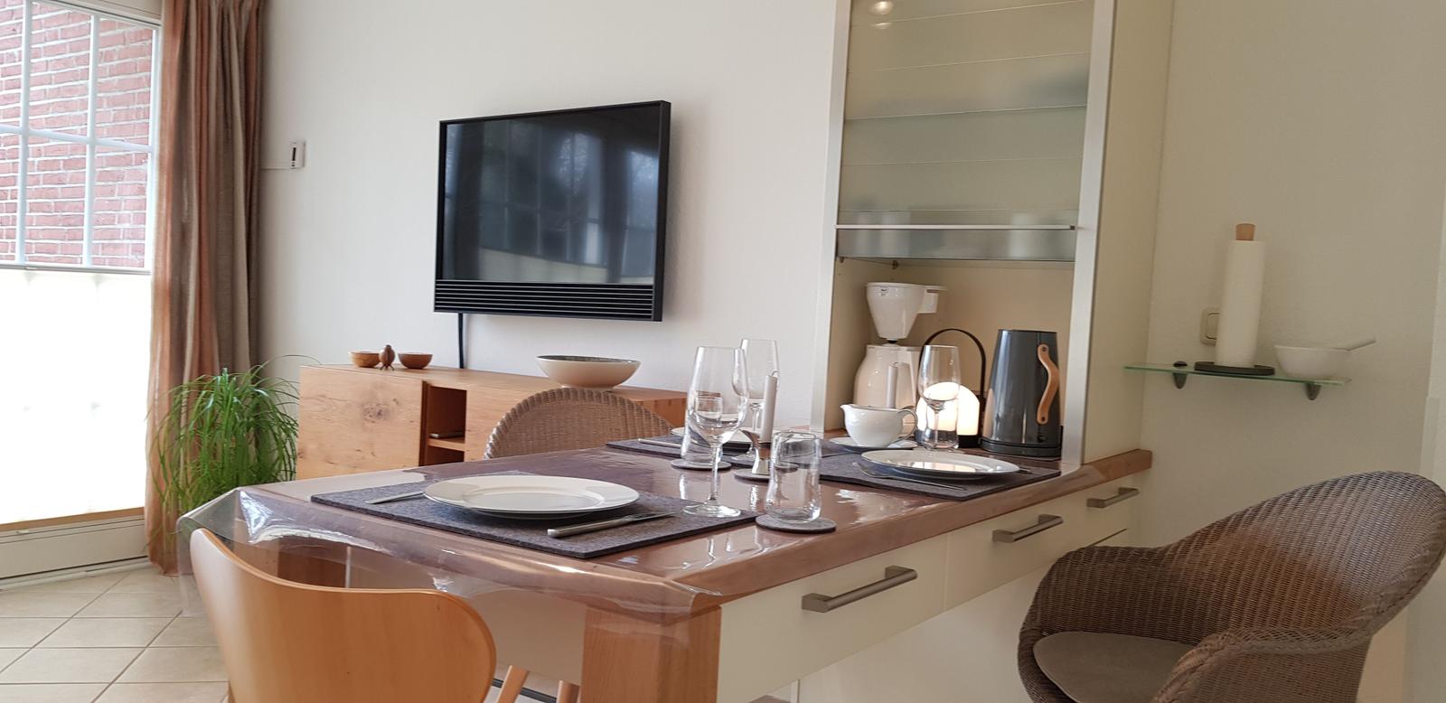 Esstisch mit Stühlen, Küchenzeile und Fernseher an der Wand.