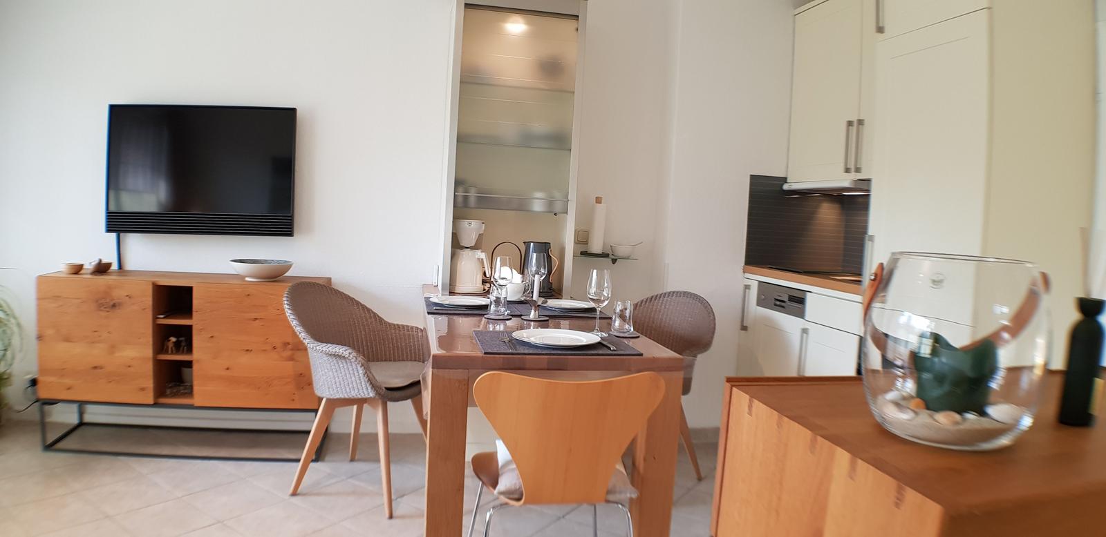 Essbereich mit Tisch, Stühlen und Fernseher. Küche mit weißen Schränken und Arbeitsplatte.