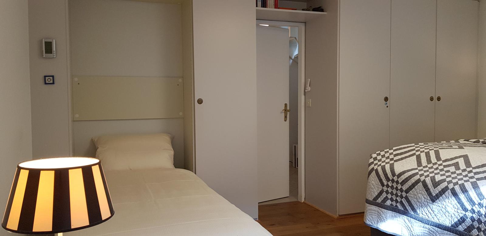 Zimmer mit zwei Betten, Kleiderschrank und Lampen. Tür führt in einen Flur.