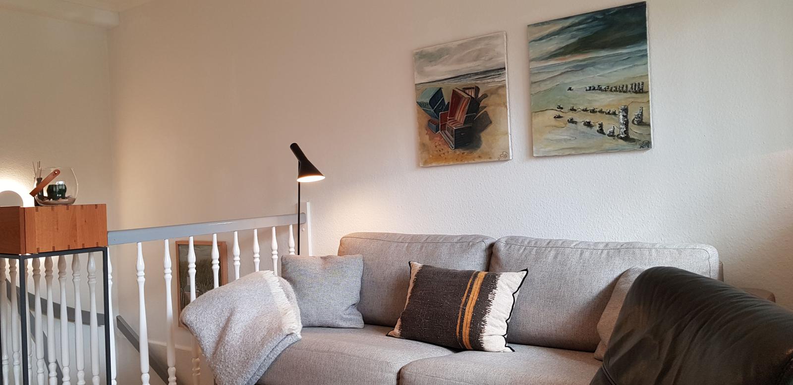 Grauer Sofa mit Kissen und Decke. Wandbilder zeigen Strand. Stehende Lampe und Holzregal.