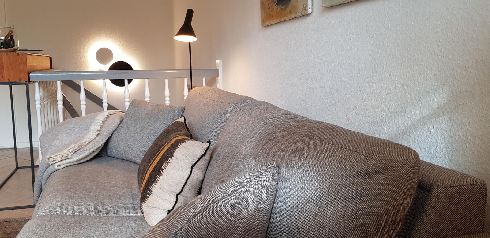 Ein grauer Sofa mit Kissen und Decke. Hintergrund mit Treppen und Lampen.