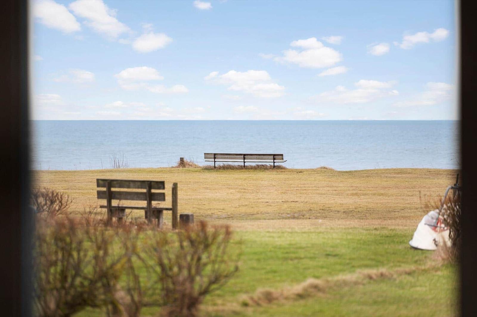 View through a window to a beach with two wooden benches and a blue sea under a clear sky.