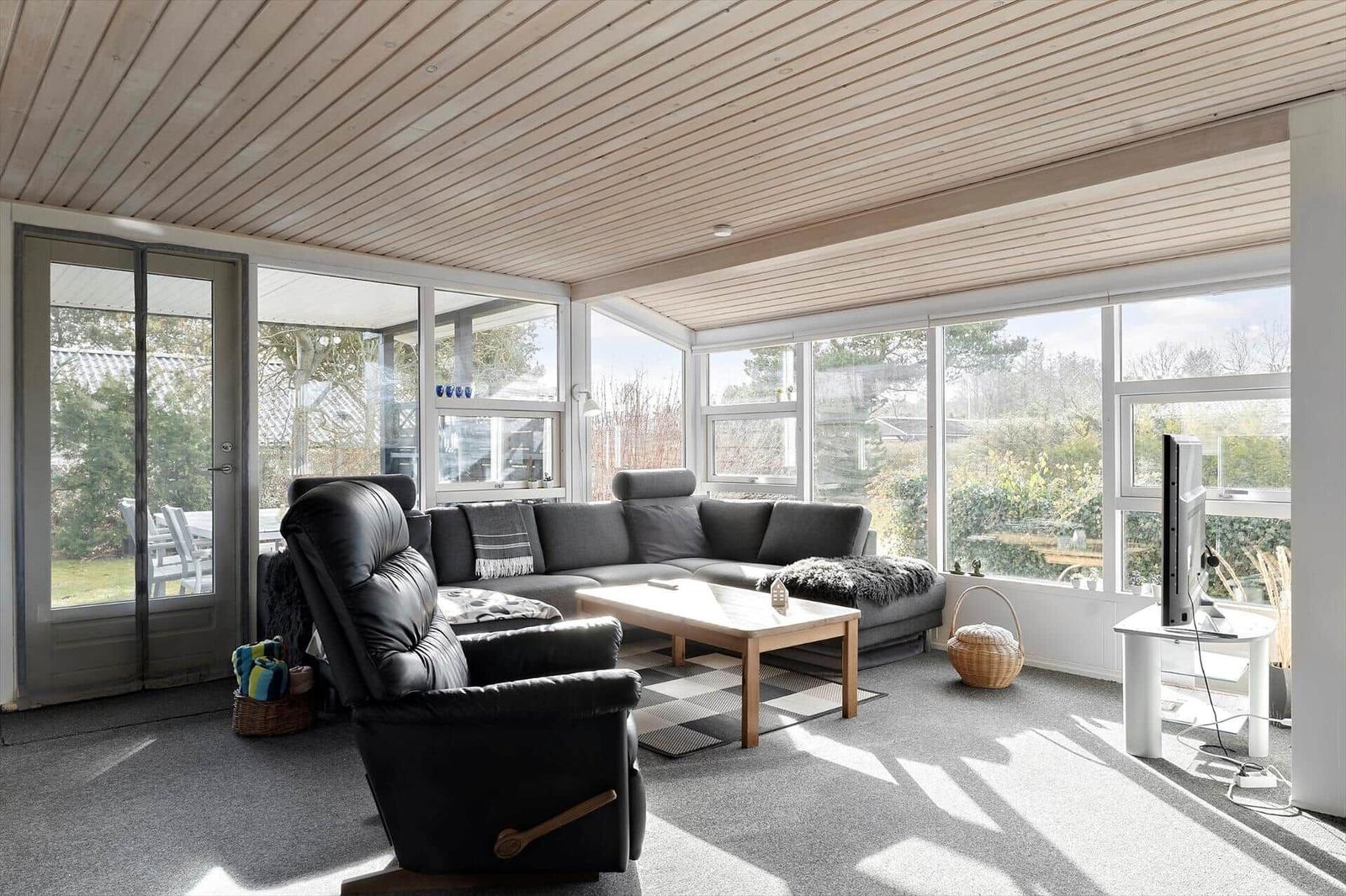 Wohnzimmer mit Sofa, Sessel und TV. Große Fenster und Holzdecke.