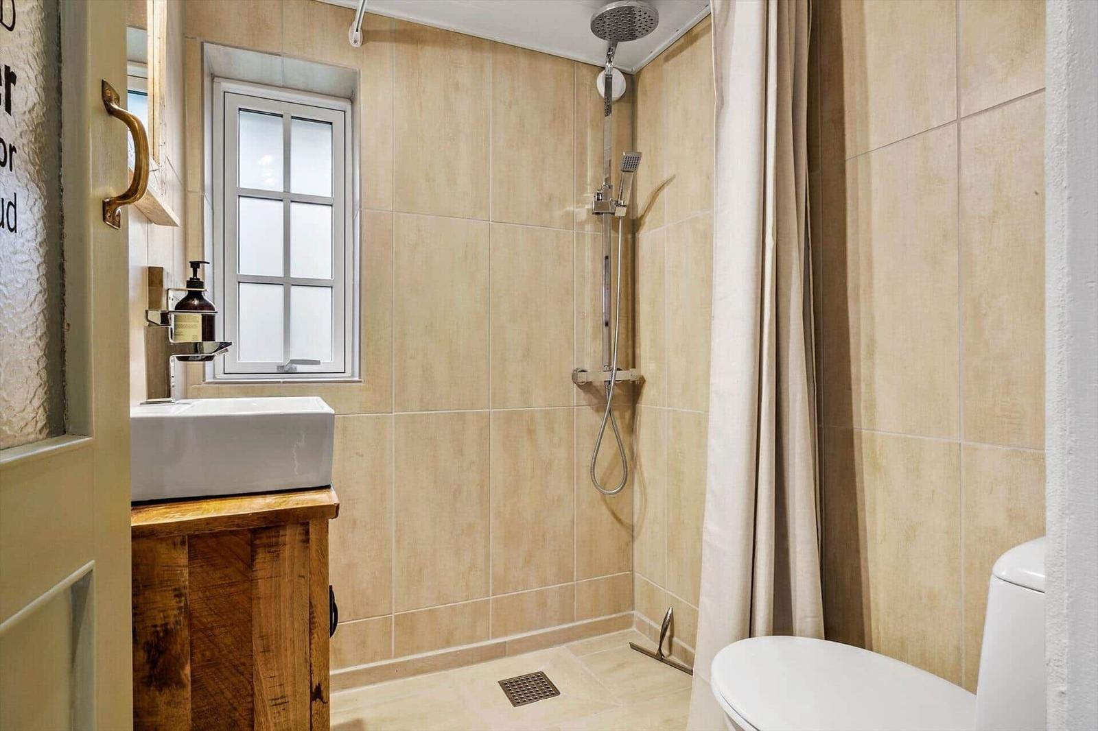 Bad mit Dusche, Waschbecken und Toilette. Fenster und Holzunterschrank sichtbar.