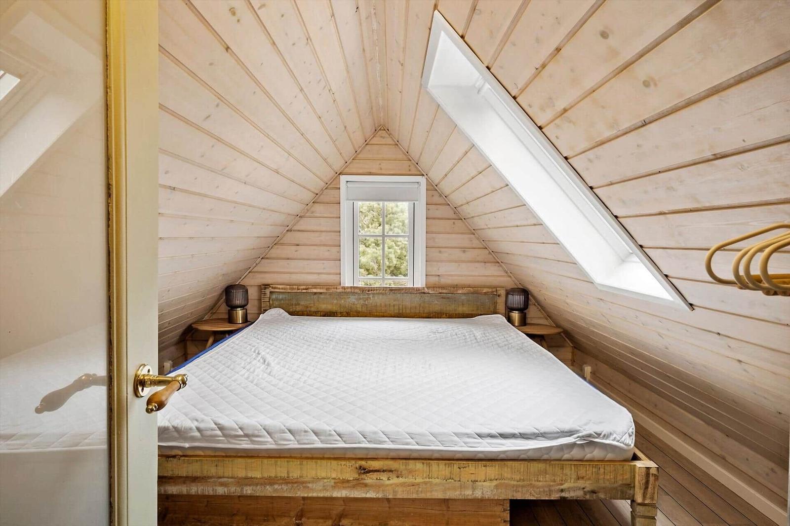Schlafzimmer mit Holzwand und Dachschrägen, Bett mit weißer Matratze, Fenster und Dachfenster.