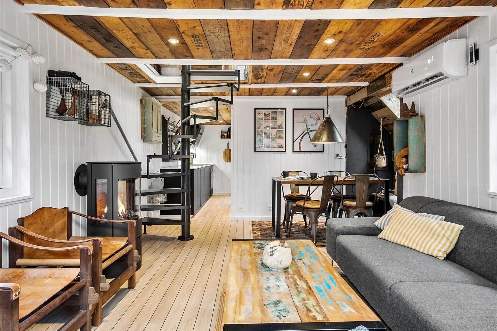 Wohnbereich mit Kamin, Sofa und Esstisch. Holzdecke und -boden. Treppenhaus im Hintergrund.