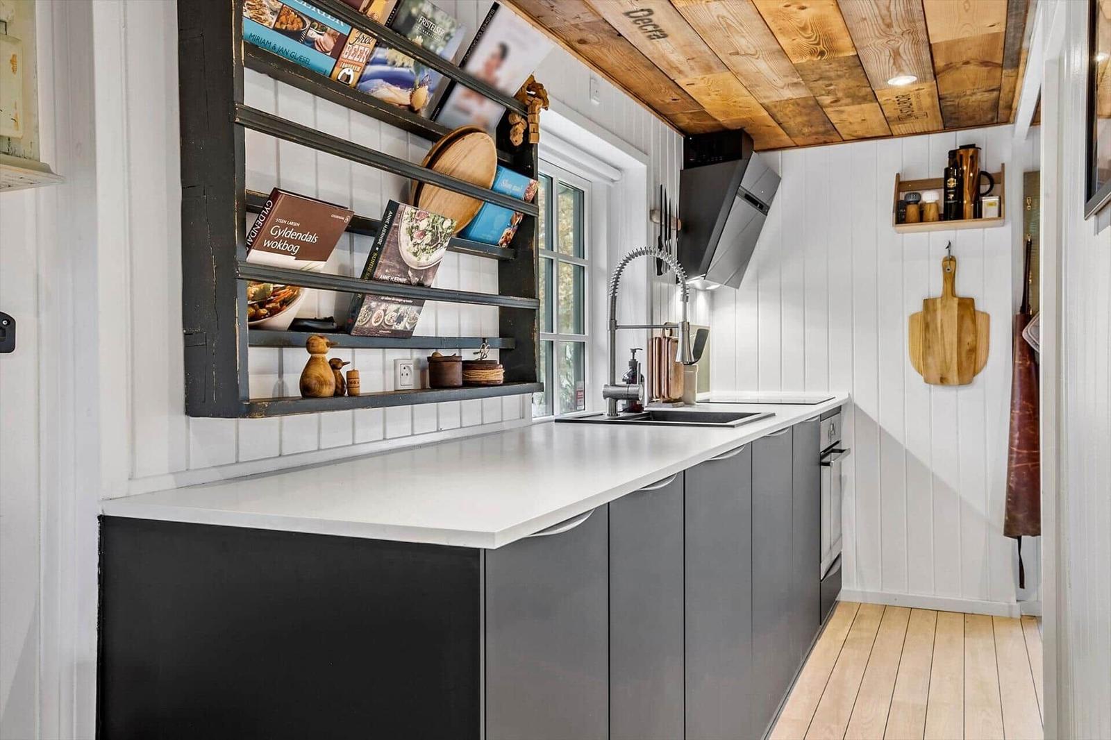 Küche mit Holzdecke, weißen Wänden und grauen Schränken. Aufbewahrungselemente und Küchengeräte sind sichtbar.