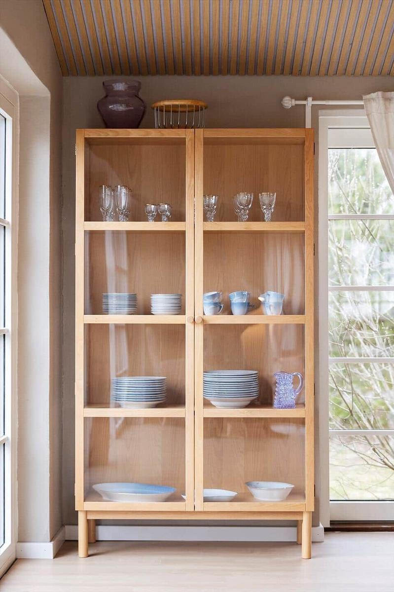 Wooden cabinet with glass doors and dishware. Windows and ceiling with wood paneling.