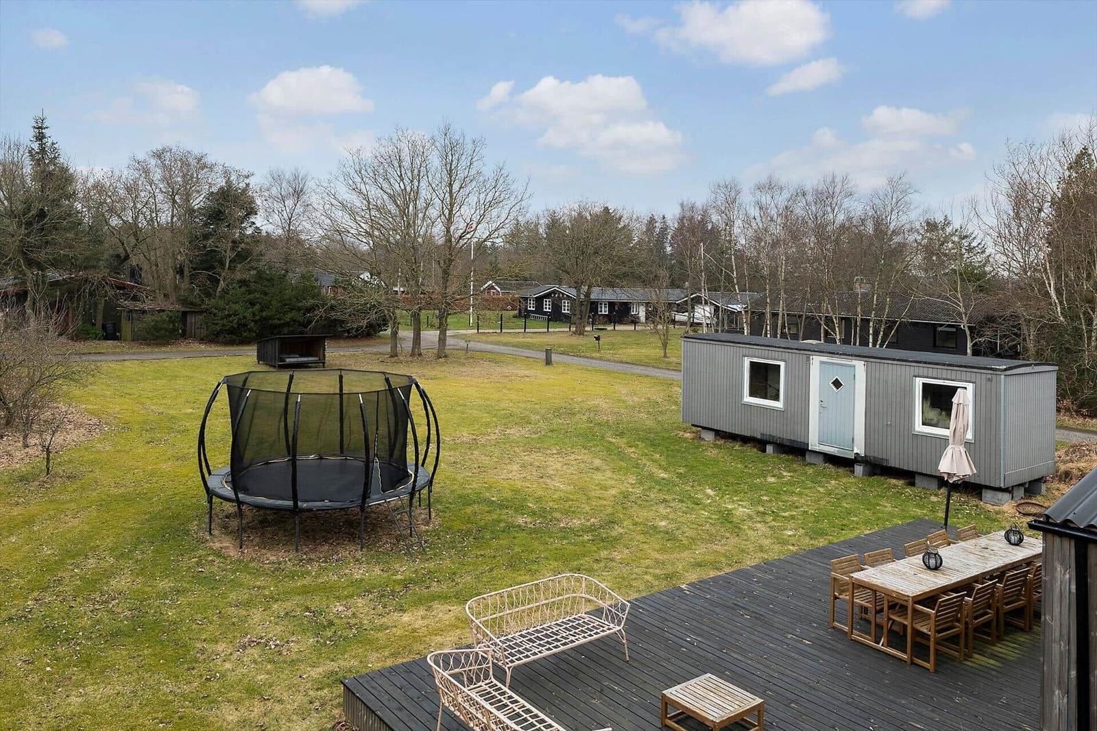 A gray holiday home with terrace, table, chairs, and trampoline on a green lawn.