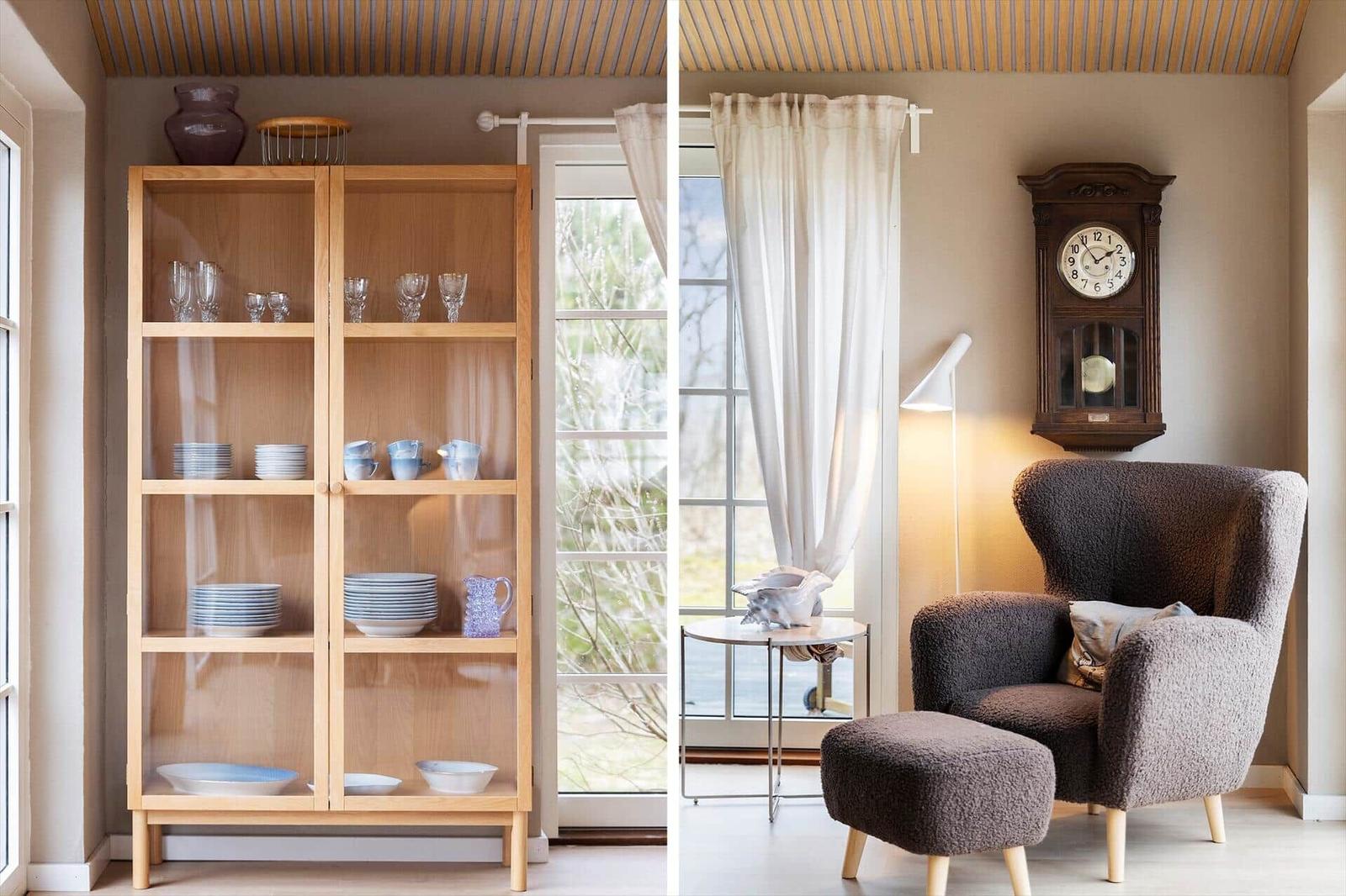 Wooden cabinet with dishes near window. Armchair with ottoman and wall clock.