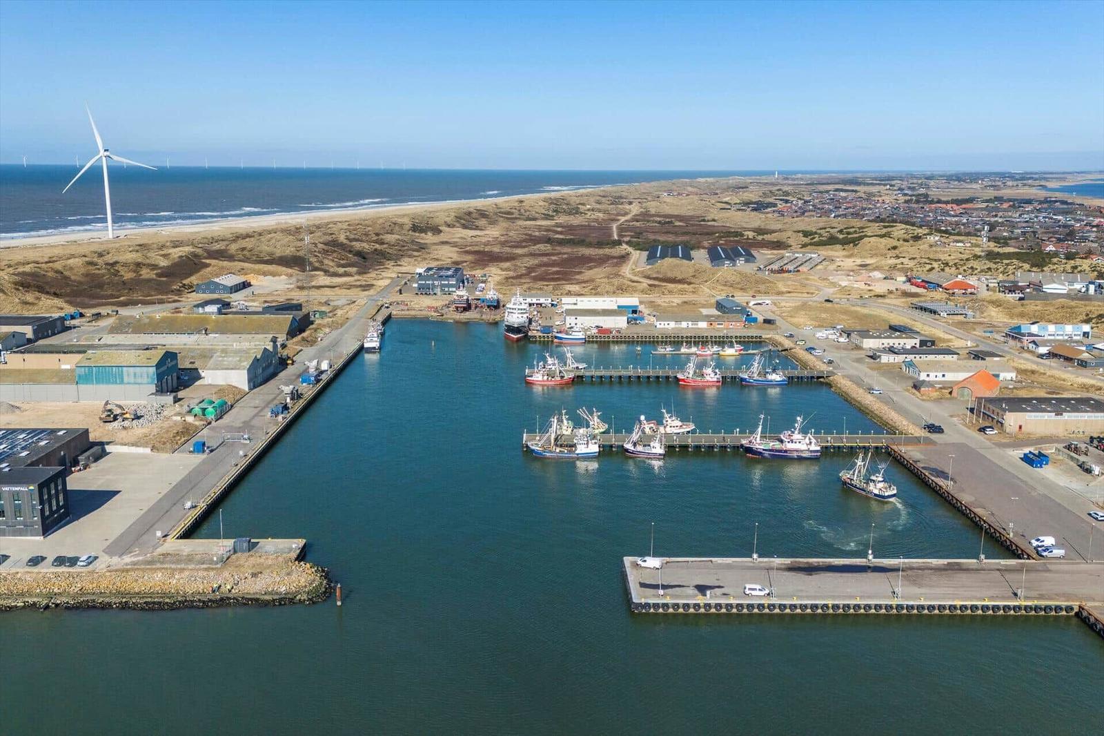Hafen mit Booten, Industriegebäude und Windkraftanlage am Strand.