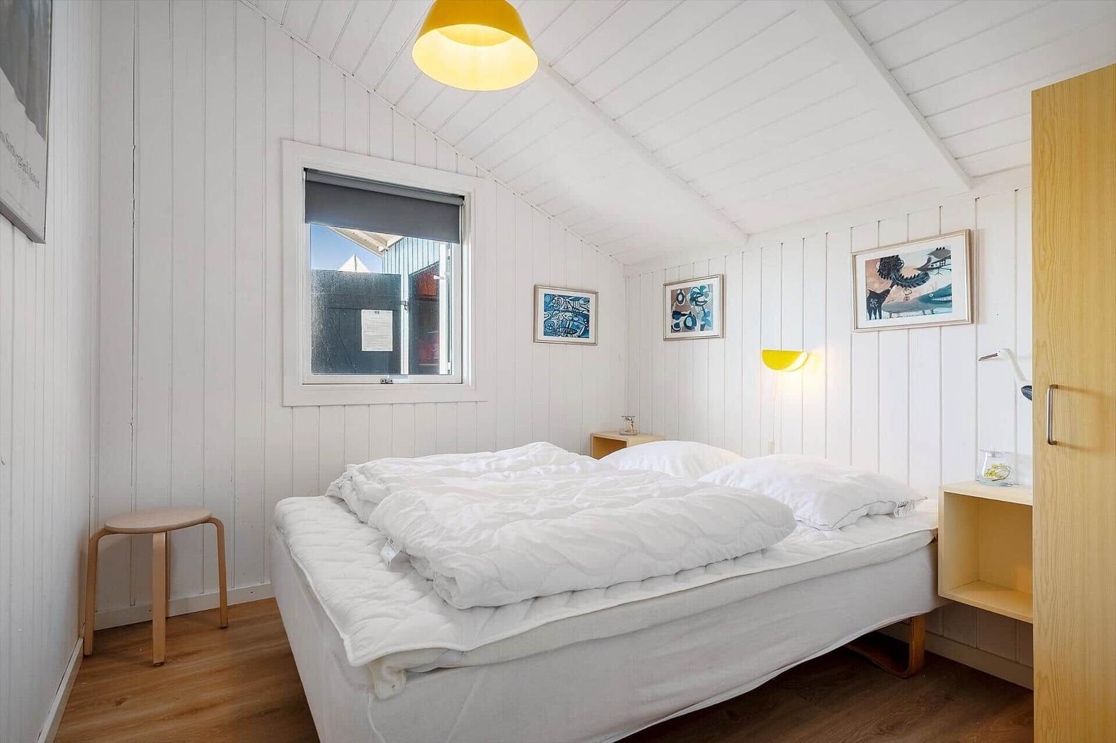 Schlafzimmer mit weißer Wandverkleidung, Bett, Fenster und gelber Lampe.