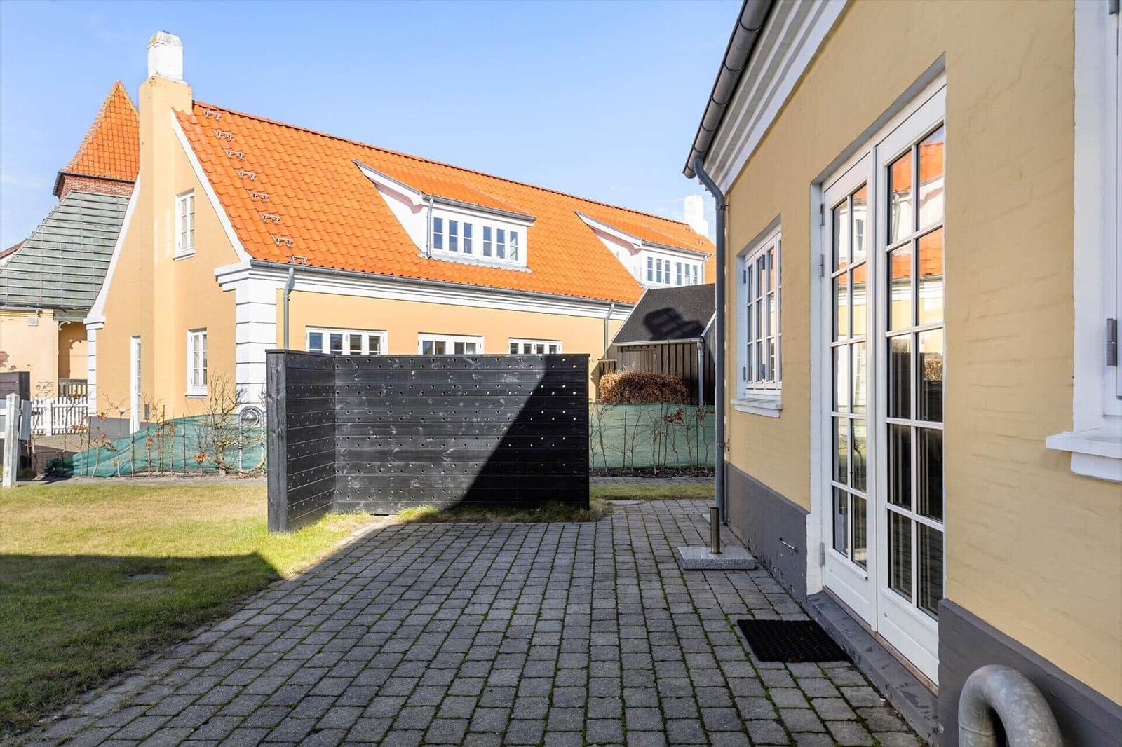 Hinterhof mit Pflasterweg, gelbem Haus und rotem Dach. Schwarze Holzwand und Fenster sichtbar.