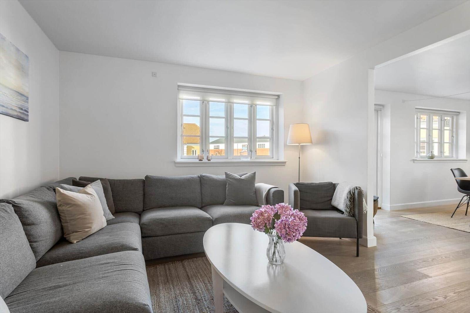 Wohnzimmer mit grauem Sofa, weißem Tisch und Blumen. Fenster zeigen Blick auf Gebäude.
