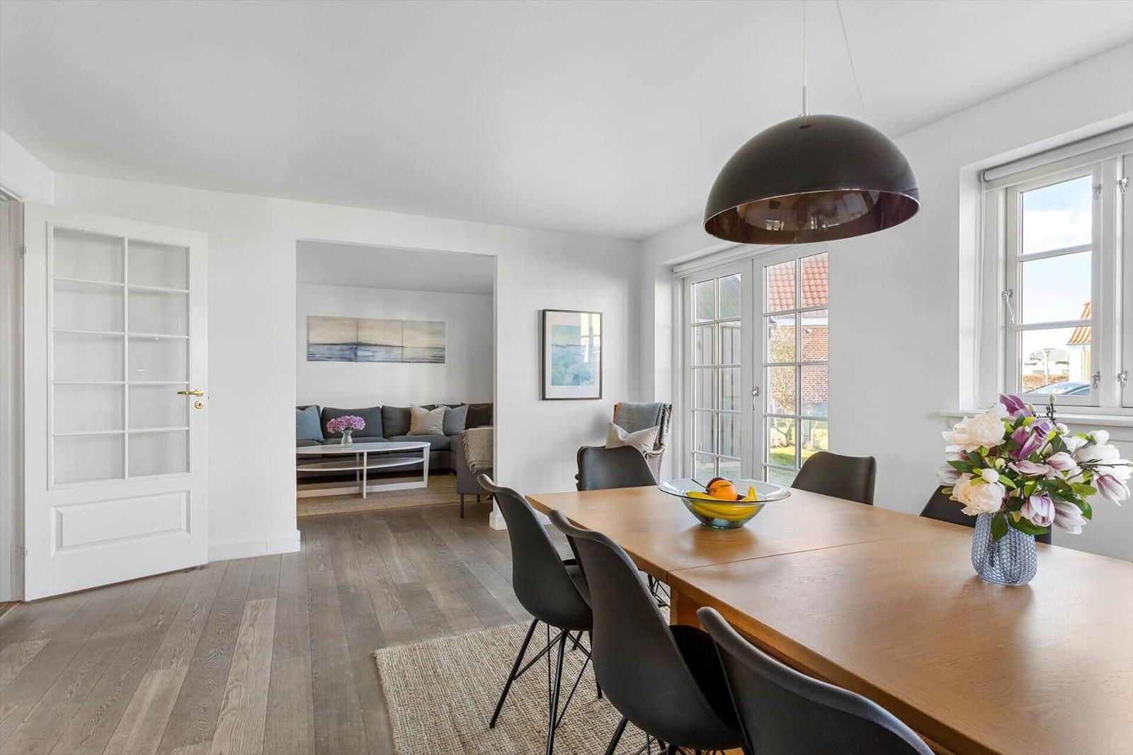 Essbereich mit Holztisch, schwarzen Stühlen und großer Hängelampe. Durchgang zum Wohnzimmer mit Sofa.