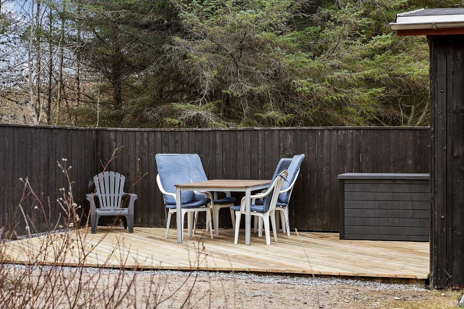 Deck with table, chairs, and wooden flooring in front of dark fence and forest.