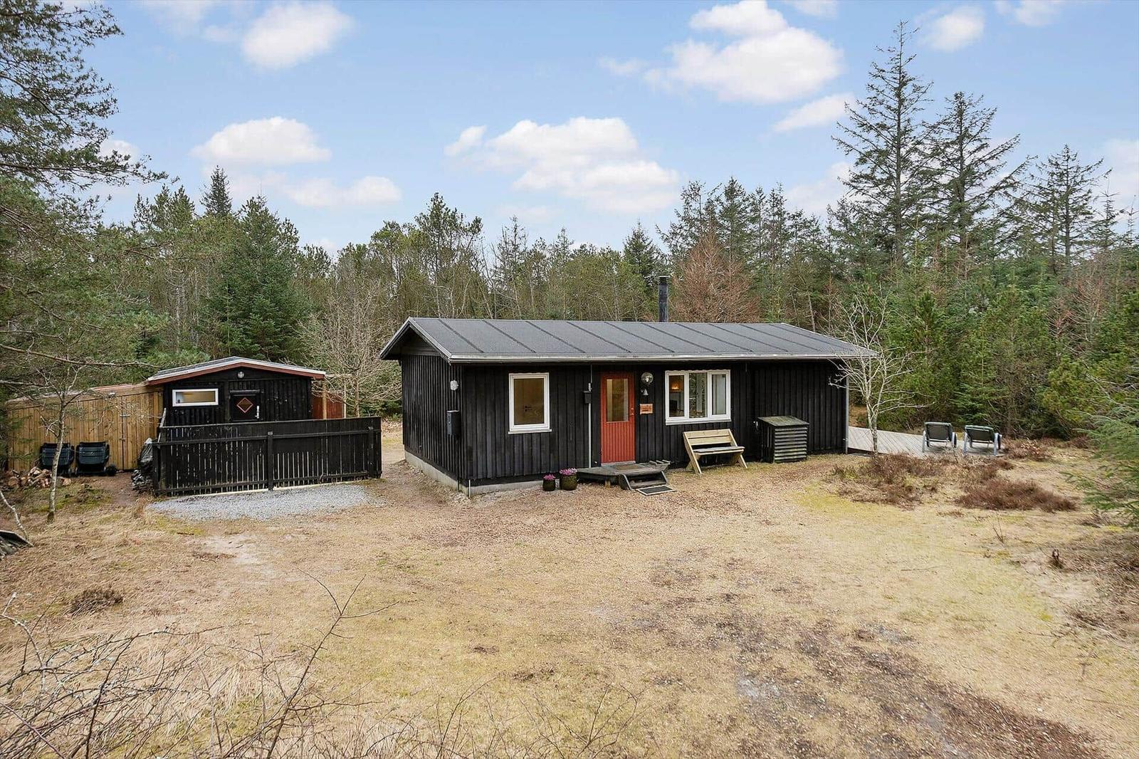 Black wooden house with red door and wooden bench in front of forest.