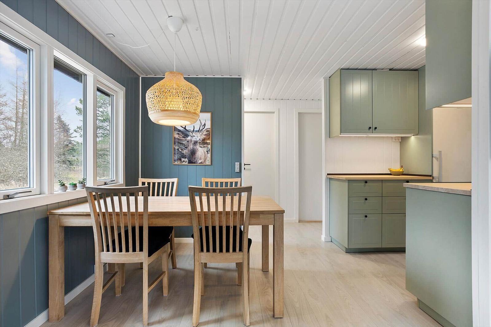 Dining area with wooden table, chairs, and kitchen. Windows with view of forest.