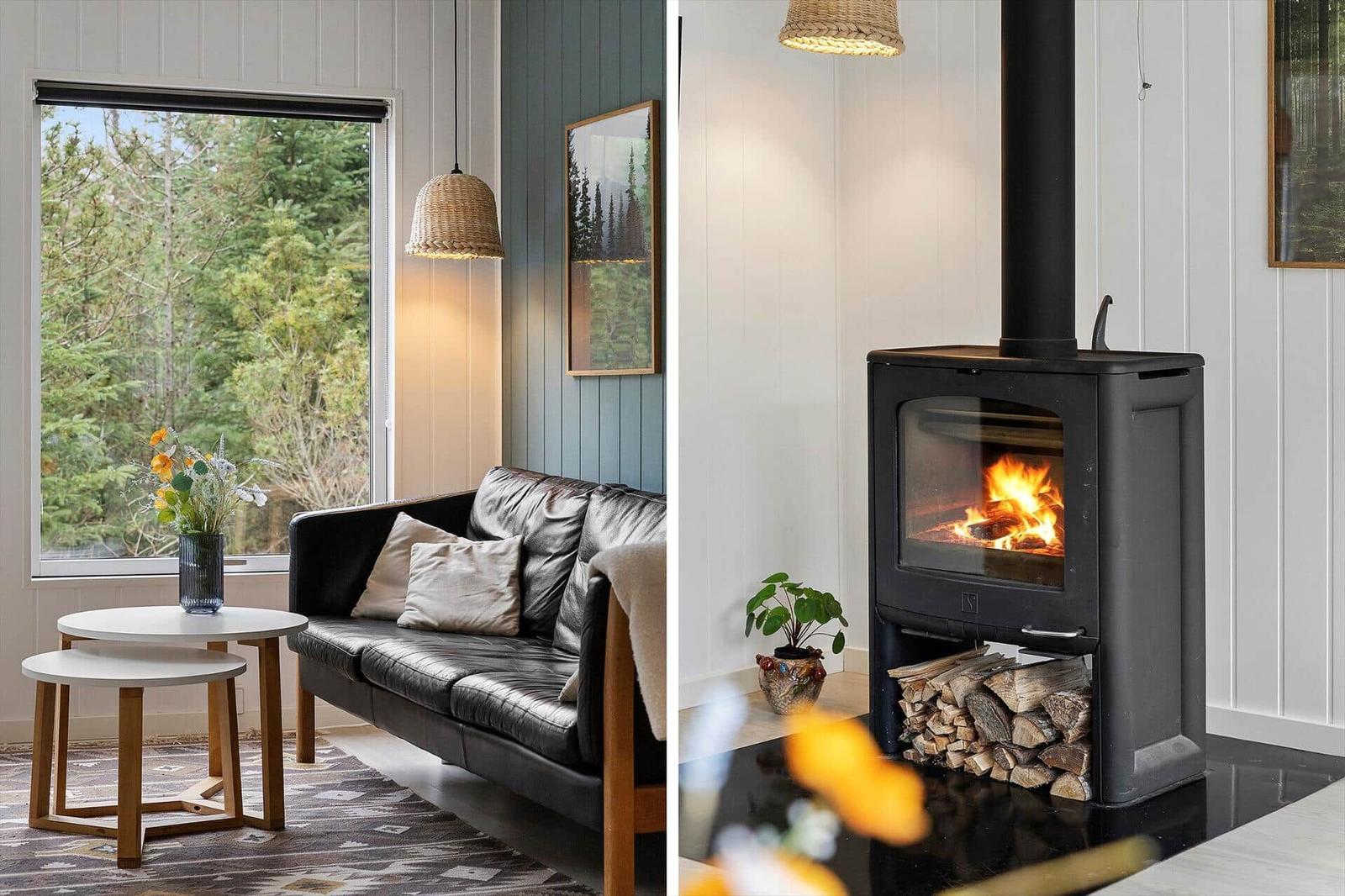 Black sofa by large window with forest view. Wood-burning stove with lit fire.