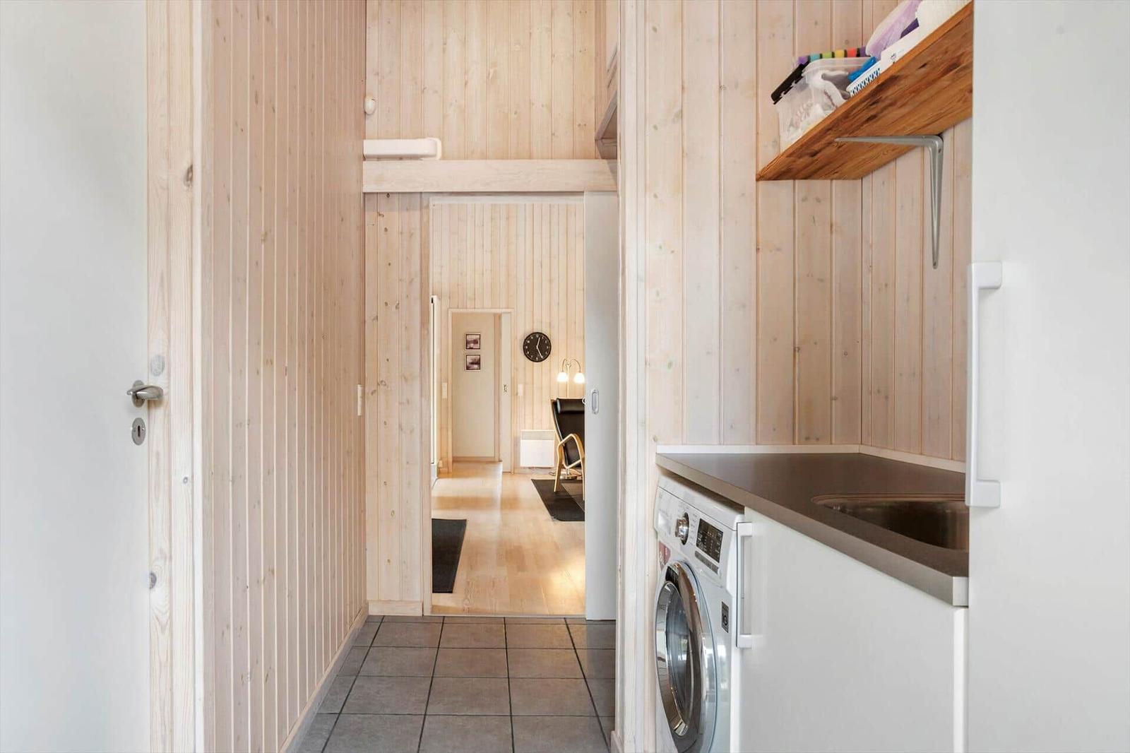 Hallway with washing machine and wooden wall, view into living area with chair and clock