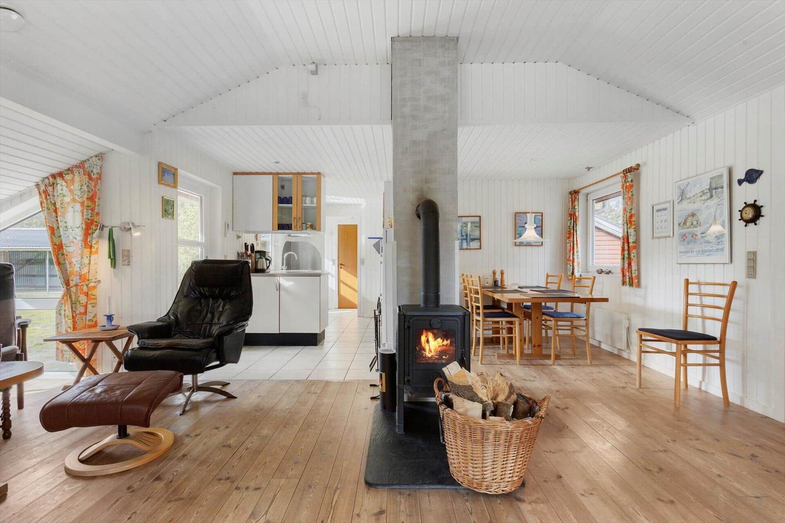 Living area with wood stove, dining table, and kitchen. Wooden floor and white walls.