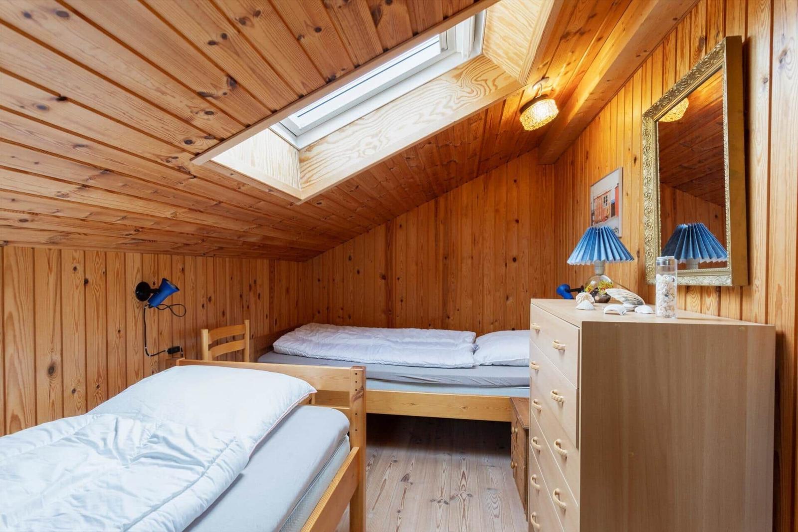 Room with two beds, wooden walls, and skylight. Dresser with mirror and lamp.