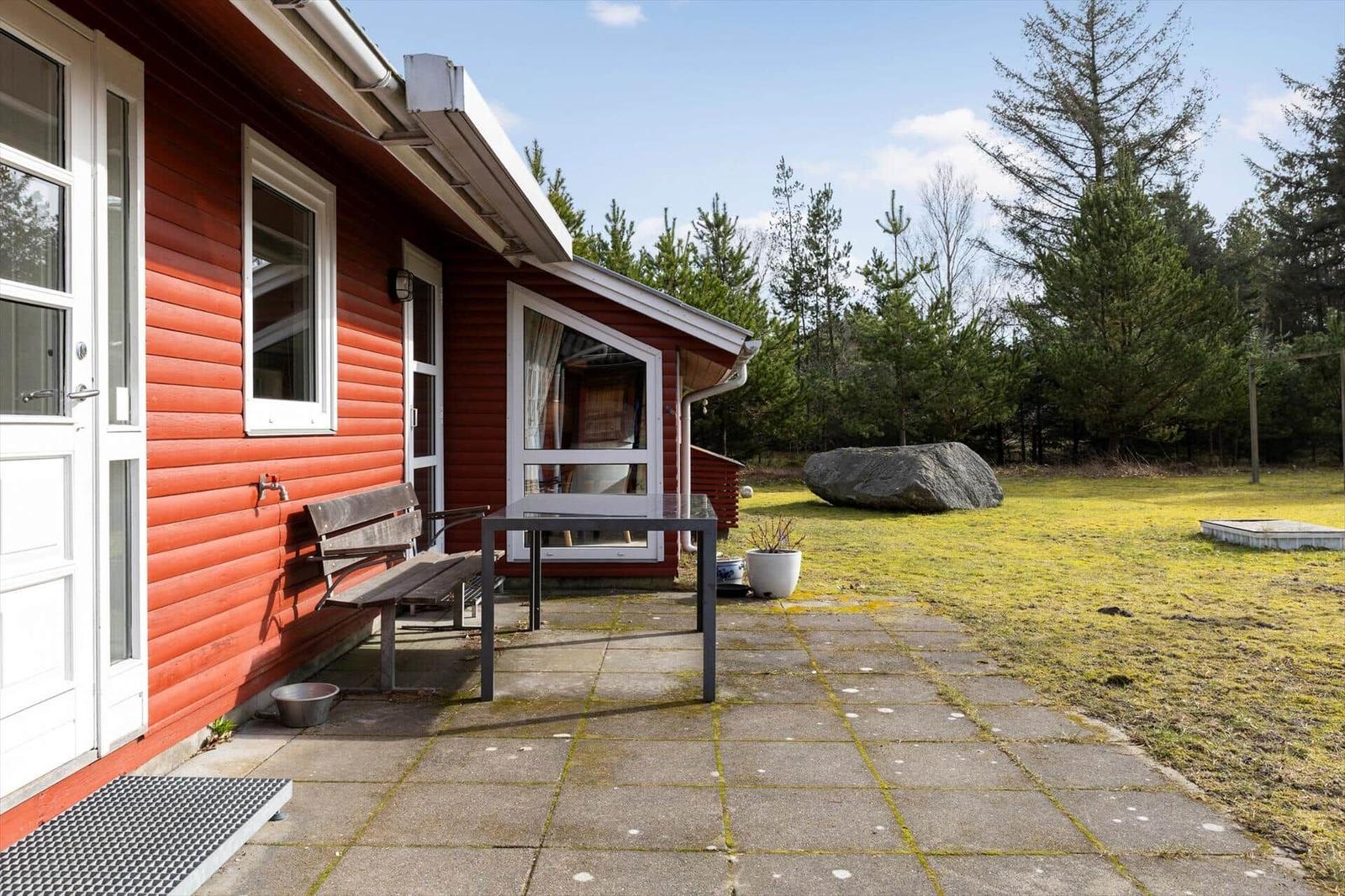Red wooden house with patio, table, bench, and garden. Forest and large stone in background.