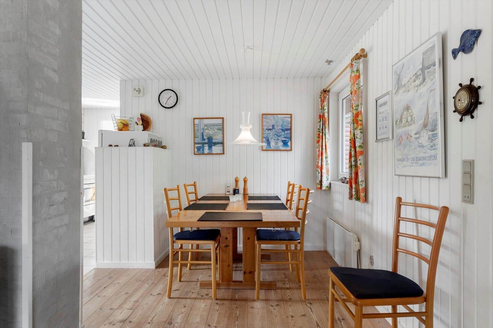 Dining room with wooden table, chairs, and white walls. Window with colorful curtains.