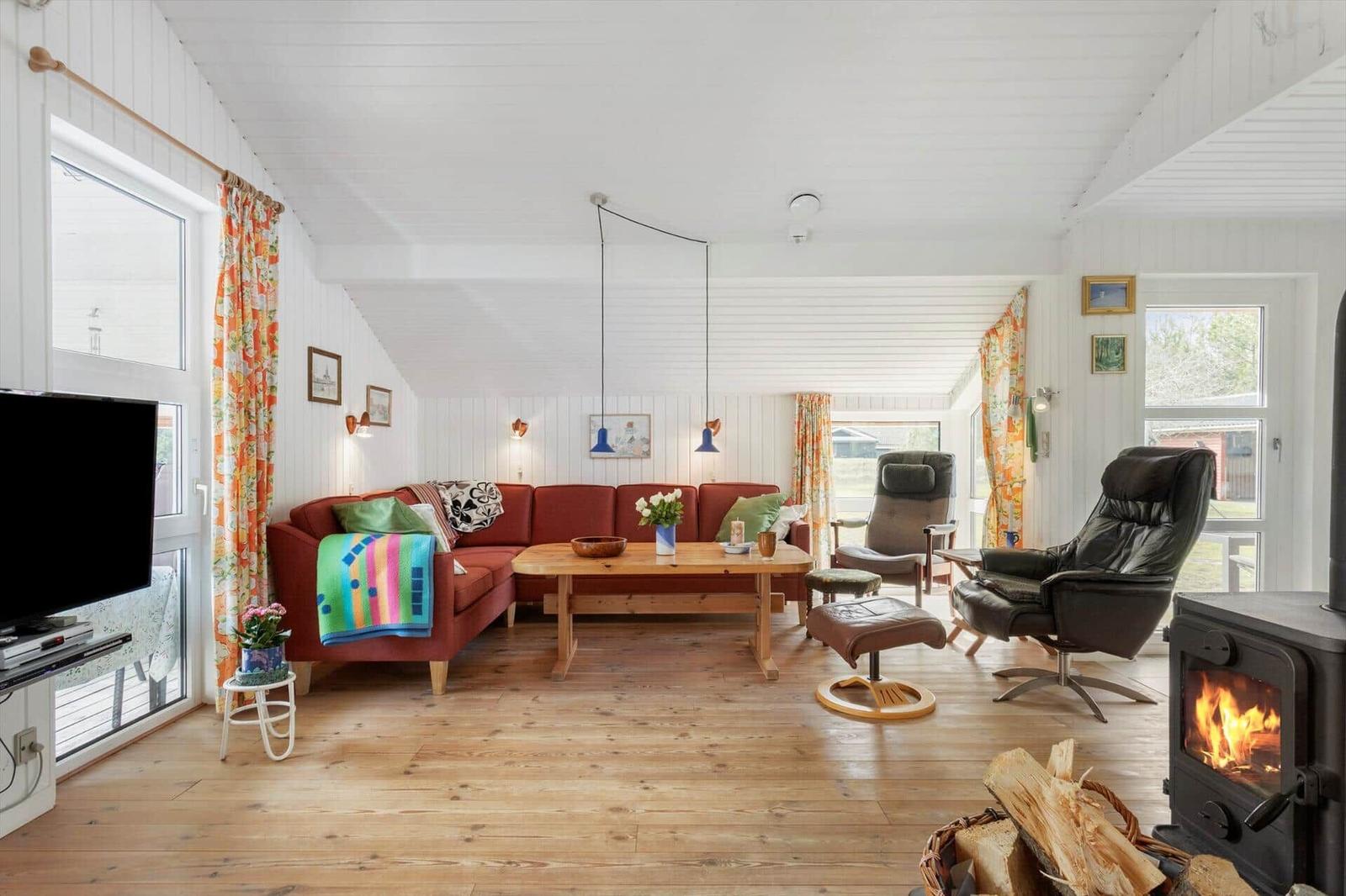 Living room with red sofa, wooden table, and wood-burning stove. Windows with colorful curtains.