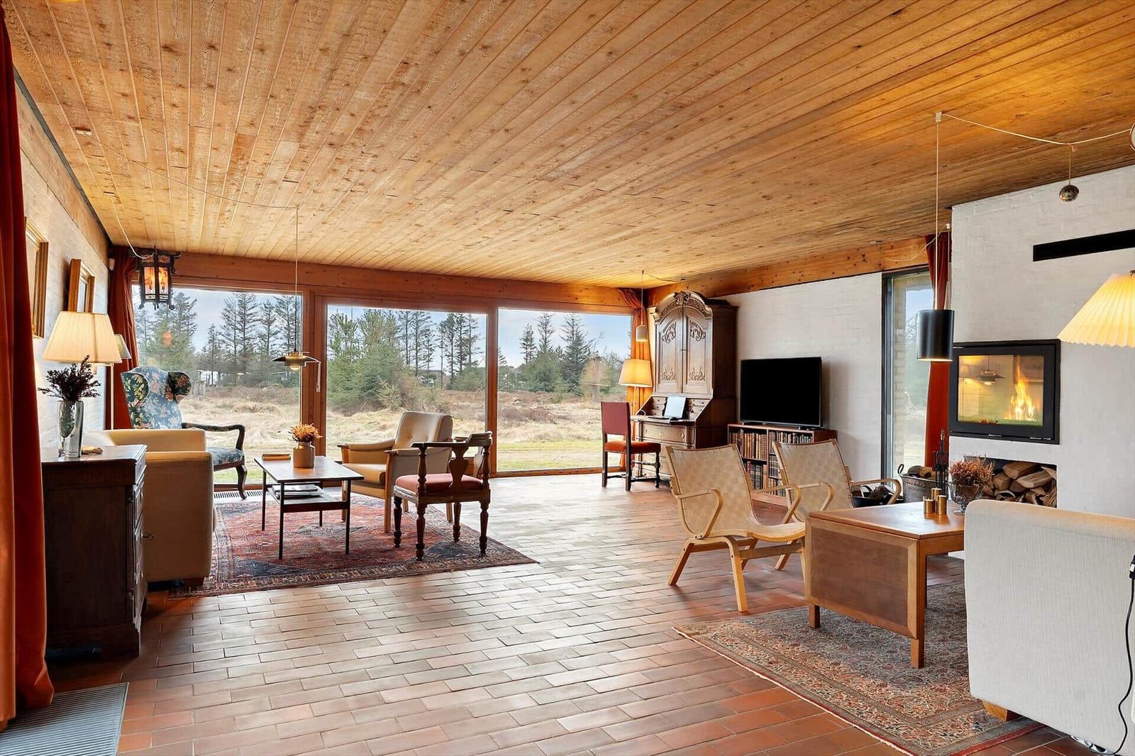 Wohnraum mit Holzdecke, Kamin und großem Fensterblick auf Wald.