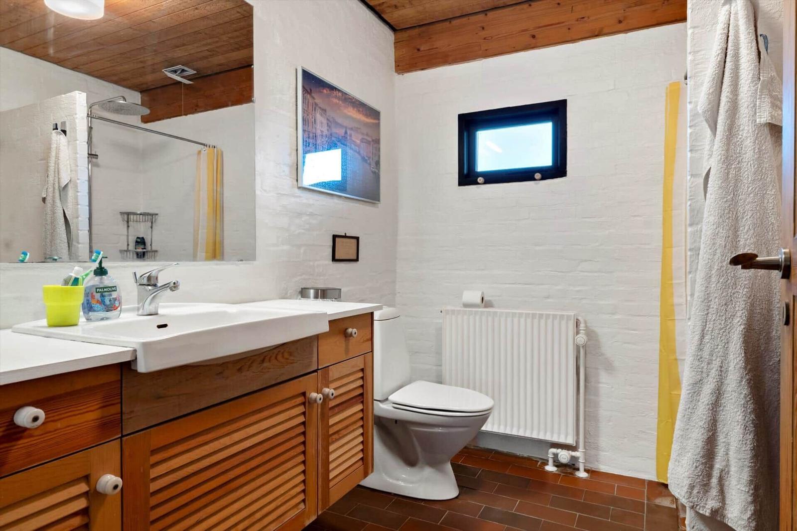 Badezimmer mit Waschbecken, Toilette und Dusche. Weiße Wände, Holzdecke und braune Fliesen.