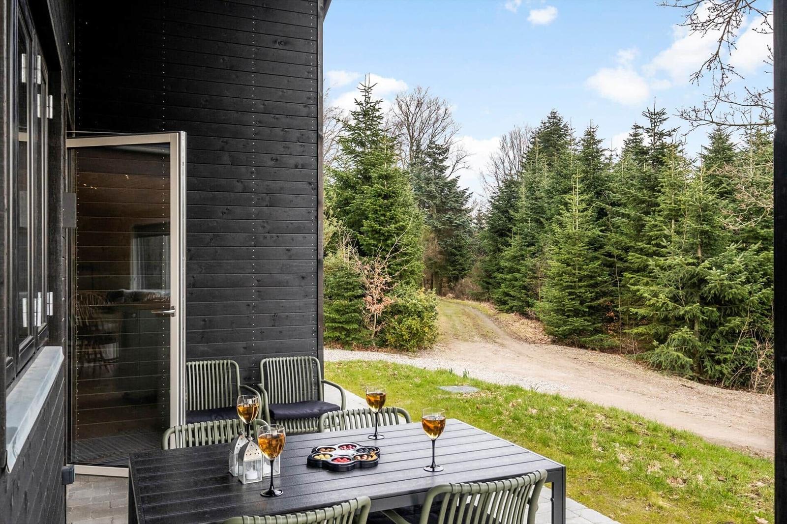 Terrace with table and chairs in front of a wooden house. View of forest and path.