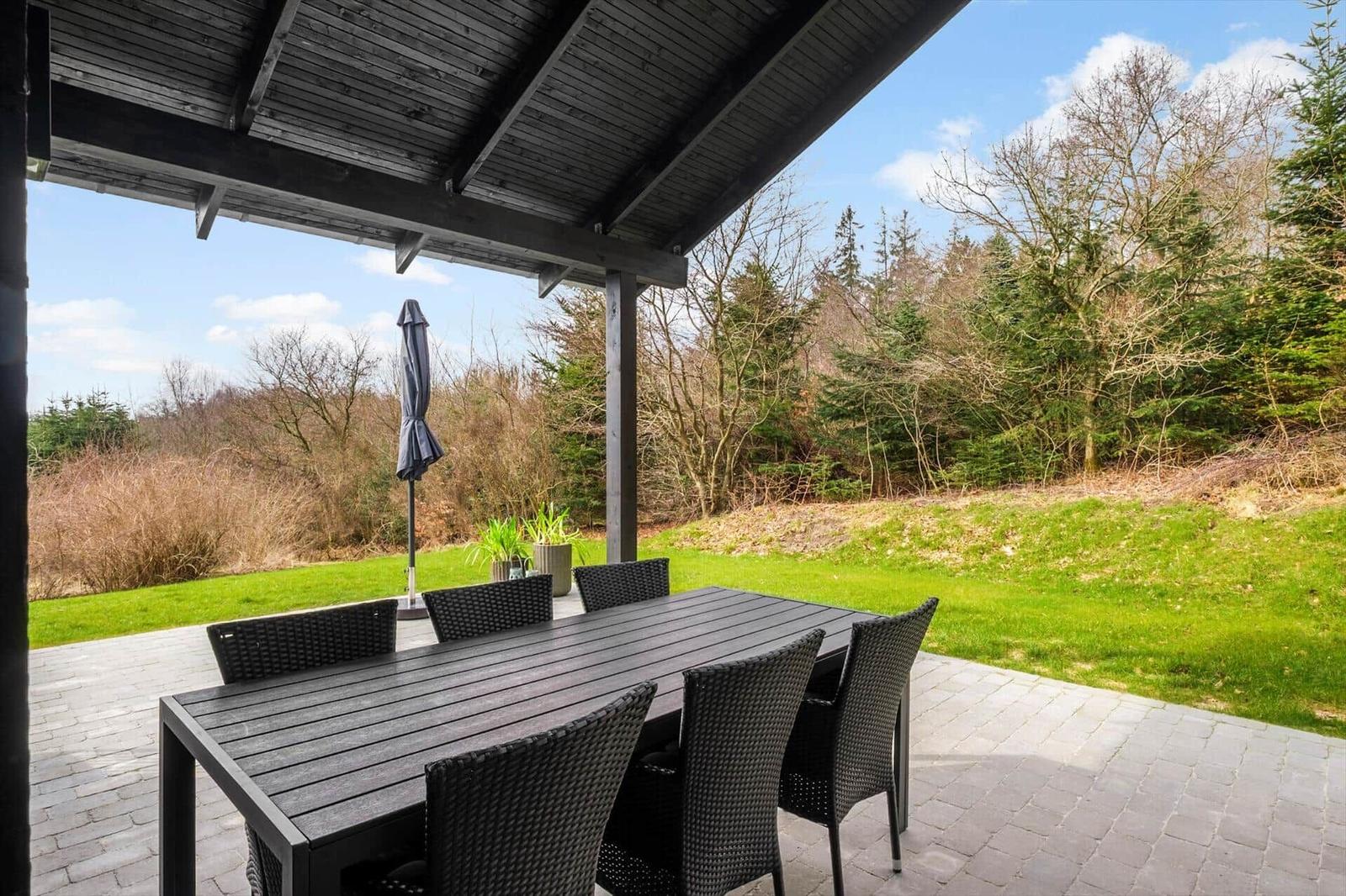 Patio with table and chairs under roof. View of green lawn and forest.