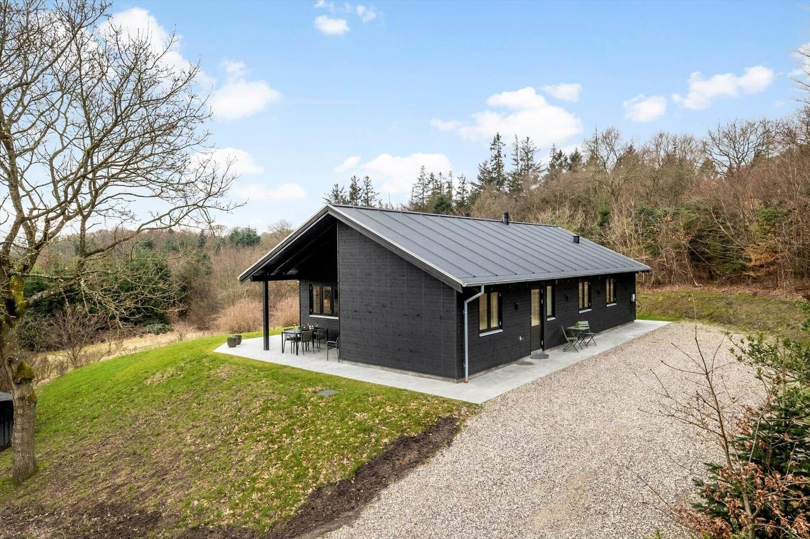 Modern black cabin with terrace, surrounded by forest and grass.