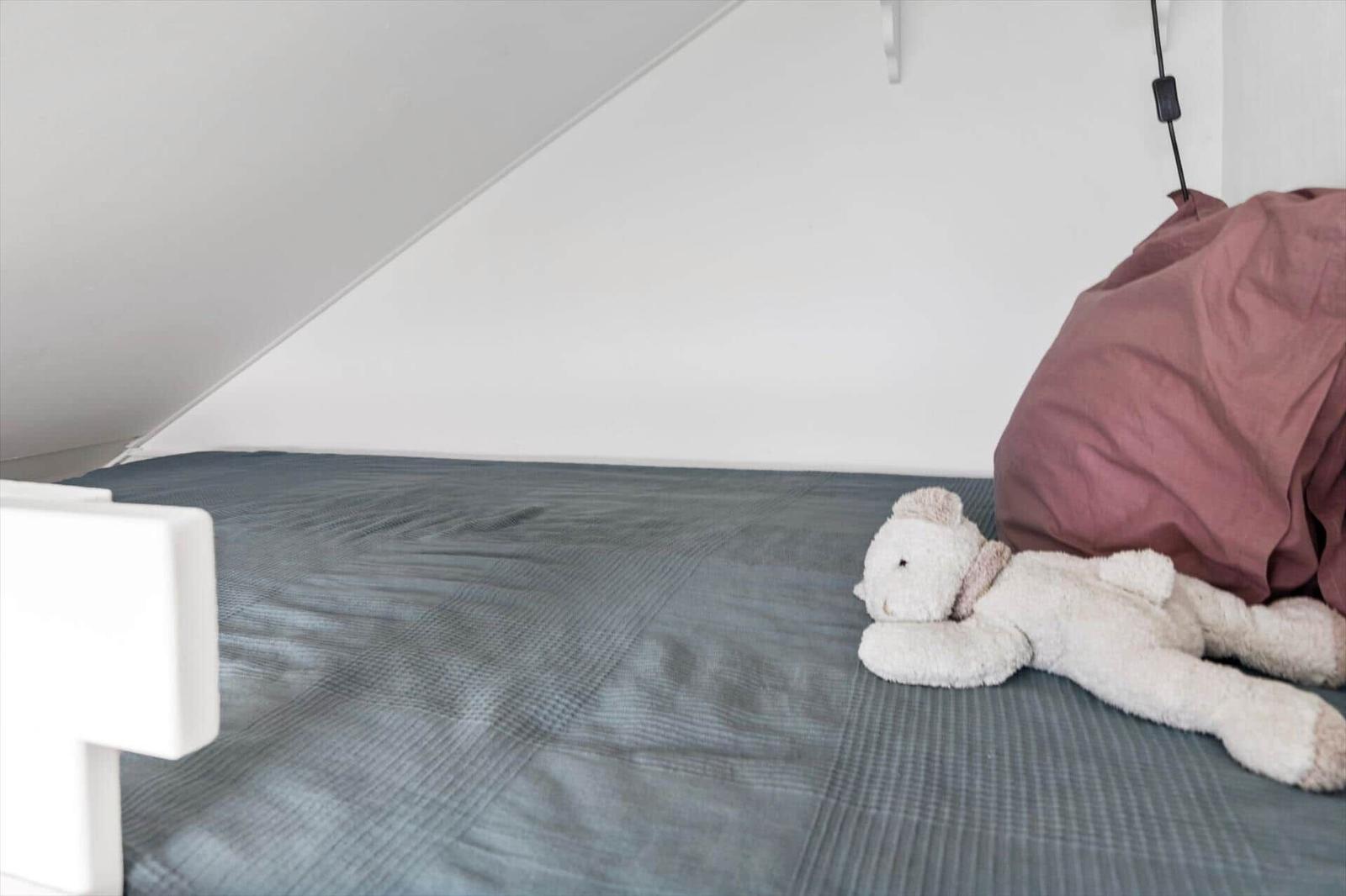 Ein Bett mit grauer Bettwäsche und einem weißen Plüschtier. Hintergrund ist weiß und schräg.