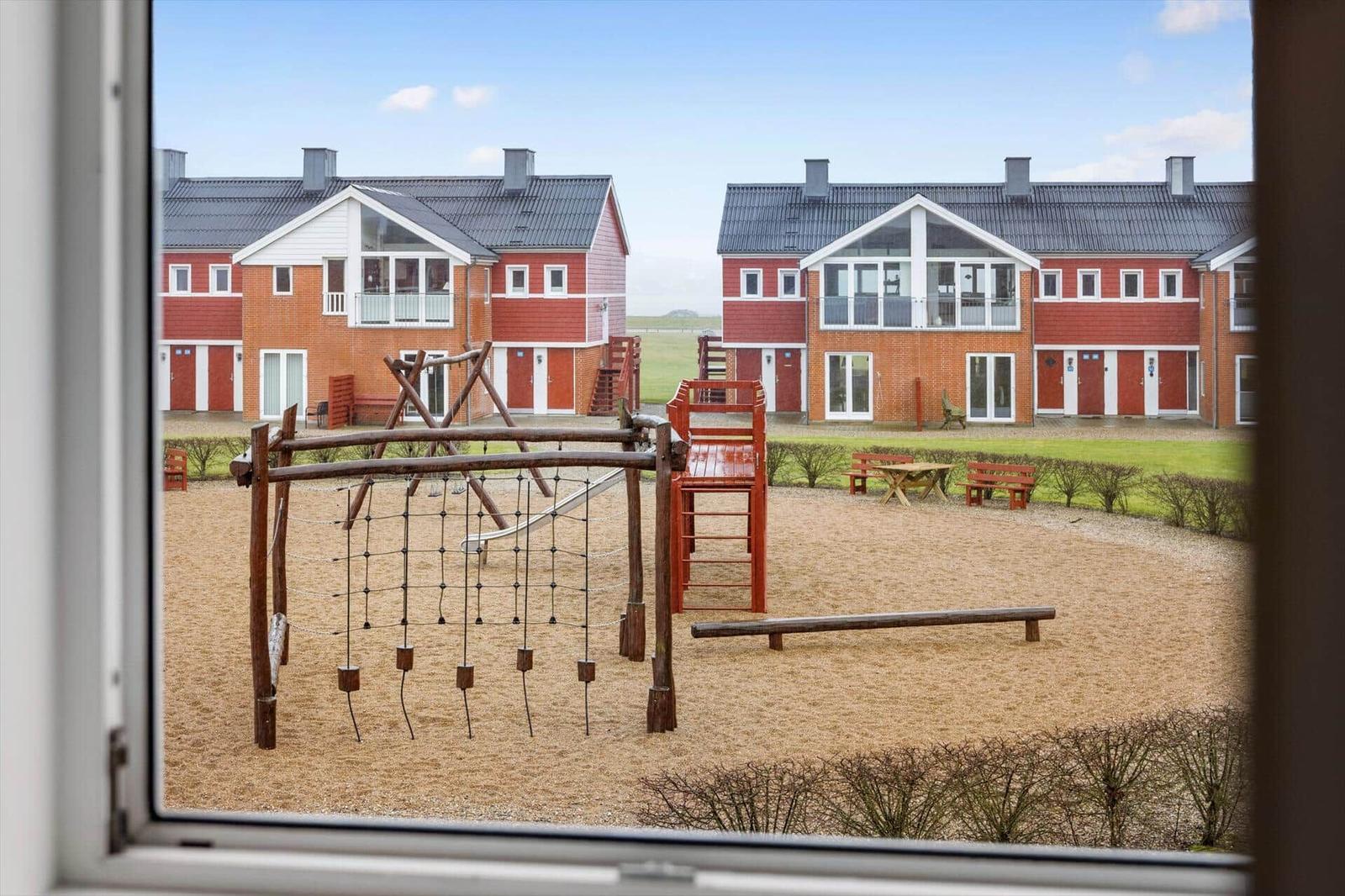 Spielplatz mit Sand und Holzgerüsten vor roten Ferienhäusern.
