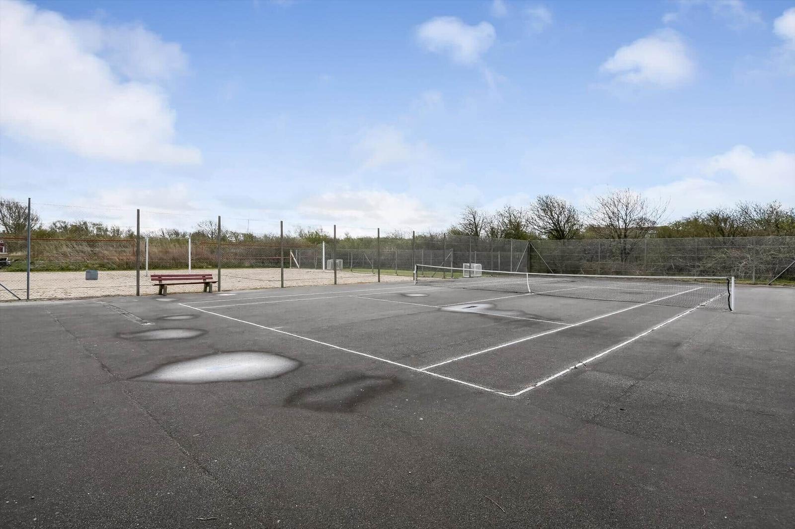 Tennisplatz mit Netz und Bank unter blauem Himmel.