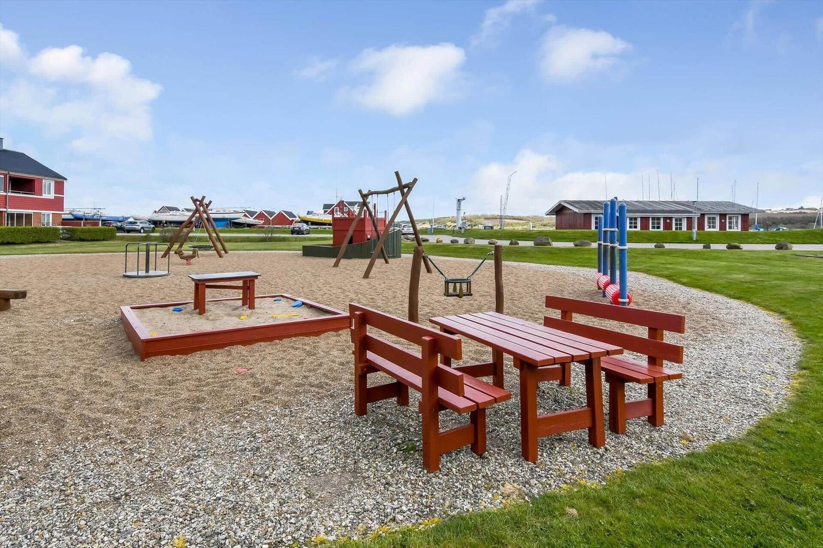 Spielplatz mit Sandkasten, Schaukel und Tischbank auf Kiesfläche.