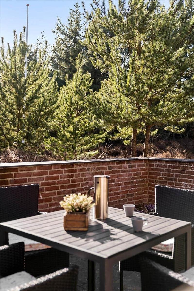 Terrasse mit Tisch, Stühlen, Thermoskanne und Pflanze. Hintergrund: Ziegelmauer und Nadelbäume.